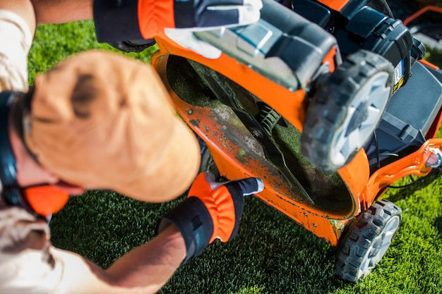 Person in gloves inspecting an orange lawn mower on grass. The mower is tilted, revealing the blades. Outdoor maintenance setting.