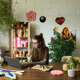 Woman with tattoos works on a laptop while holding a mug. Yarn and tufting supplies on table. Creative studio with colourful decor. Calm mood.