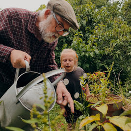 Elderly man in a plaid shirt and child garden together, watering plants with a can. Lush greenery in the background, creating a peaceful mood.
