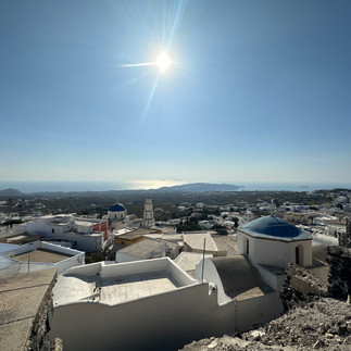 view of the top of homes in Pyrgos Santorini Greece with the sun shining over the ocean in the distance