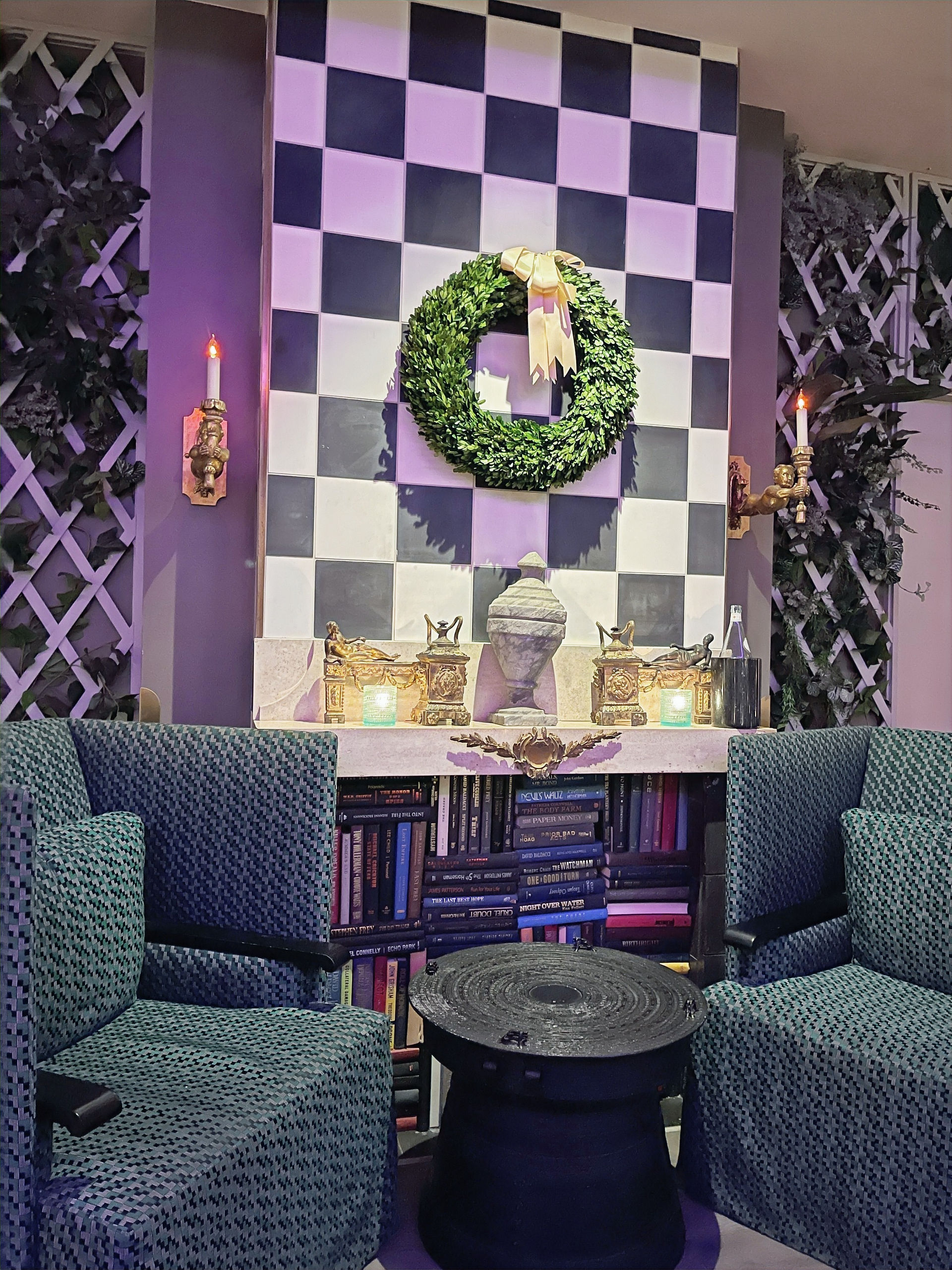 Two chairs beside a mantle with books below it.  White trellises with greenery in them behind the chairs 