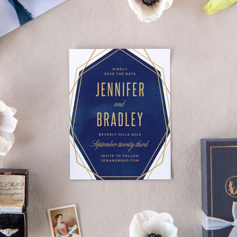 wedding flat lay of a white and navy save the date card with gold geometric lines, loose flowers and a ring in a ring box