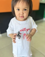 A toddler wears a Brownsville Piano Studio shirt with a treble clef and heart design.