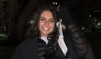 Smiling woman graduate in dark graduation gown and cap against dark backgroud.