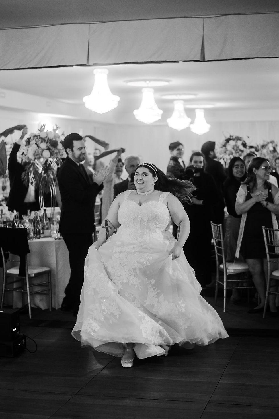 Bride in a lace gown joyfully walks through clapping guests at a wedding hall with chandeliers, creating a festive and celebratory mood.