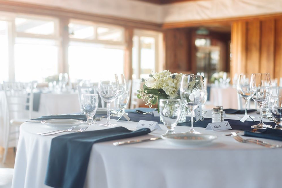 Elegant dining table setup with white cloth, navy napkins, glassware, and a flower centerpiece. Warm wooden interior in the background.