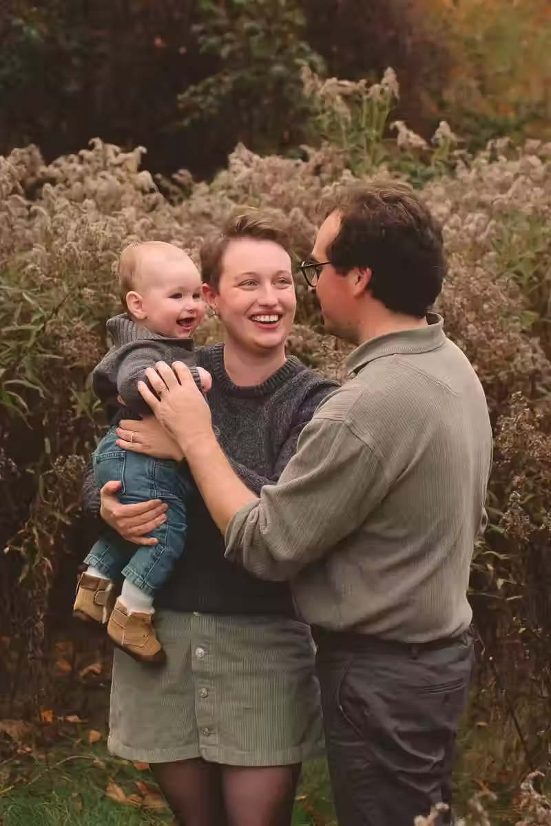 A young mother and father standing in a golden field with their laughing toddler in their arms.