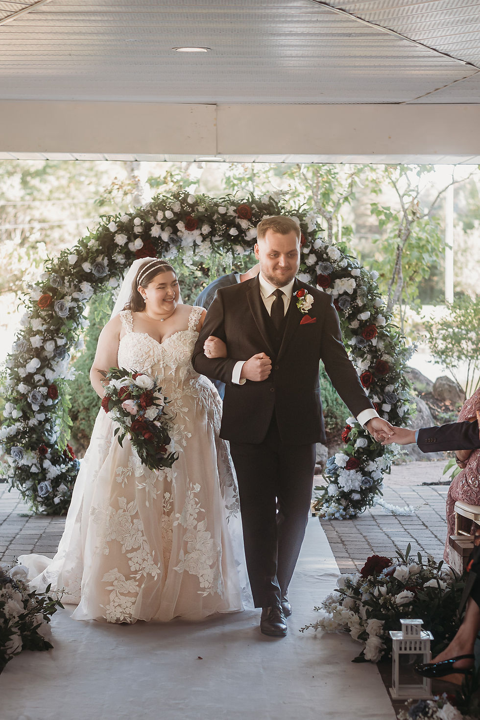 Bride and groom walking arm in arm under a floral archway. The bride holds a bouquet, both smiling. Outdoors setting with white flowers.