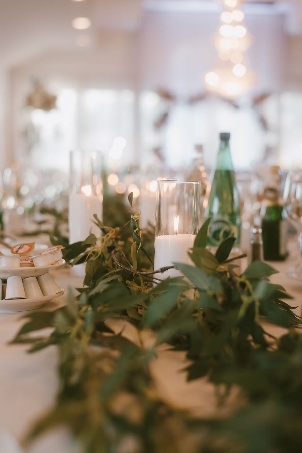 Candles in glass holders surrounded by greenery on a white table. Soft lighting, bottles, and blurred elegant background create a serene mood.