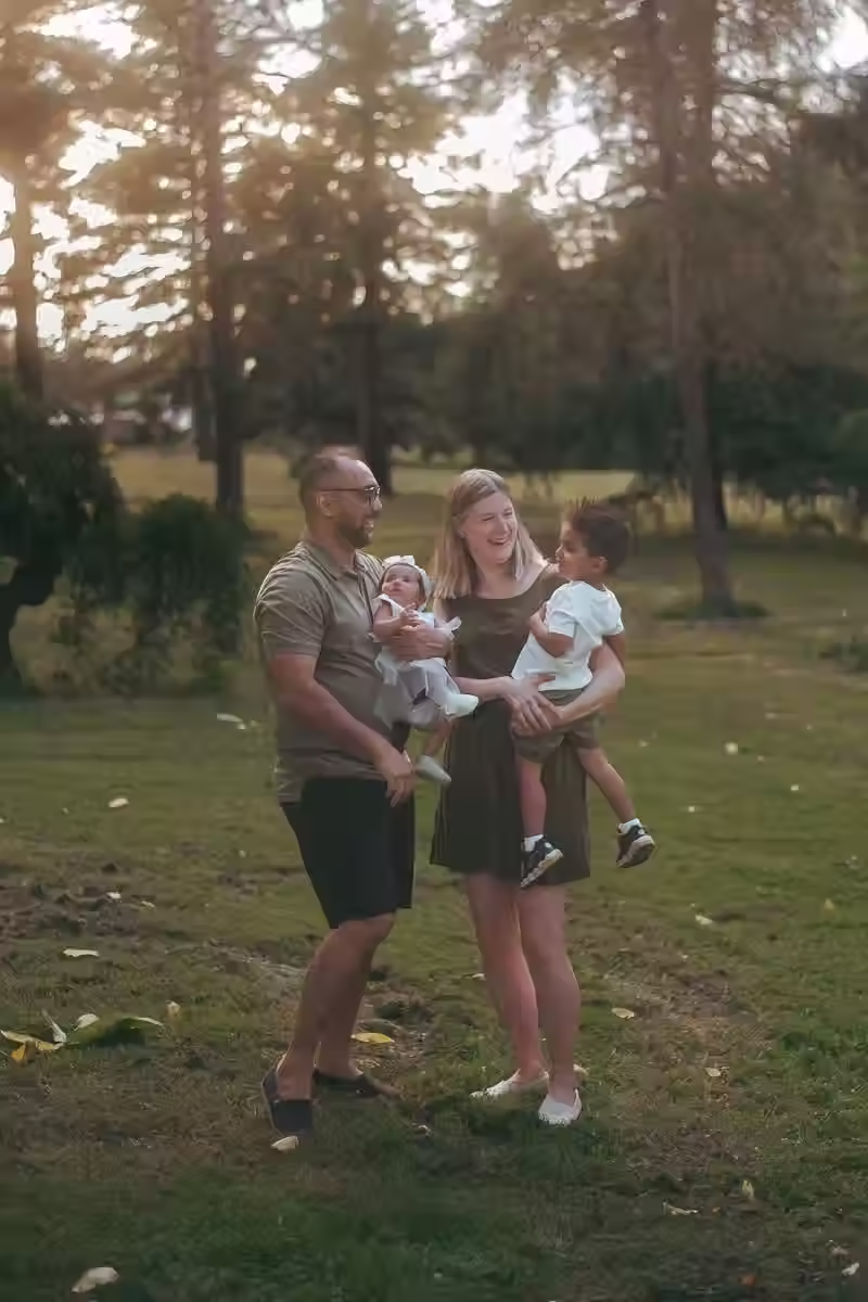 A couple tickling and laughing with their two young children in a park setting during golden hour.