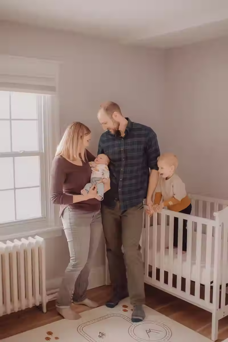 Lifestyle newborn photography in Ottawa: parents holding their infant while their young son stands in a white crib, captured in a moody, film-like indoor setting with soft natural window light.