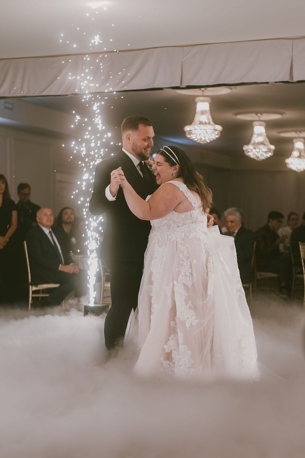 Bride and groom share a joyful dance amid fog and sparklers, with guests seated under elegant chandeliers in a celebratory atmosphere.