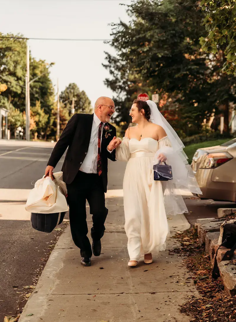 Bride in white dress and veil walks with an older man in a suit, smiling. They hold hands on a leafy street. He carries a bag.