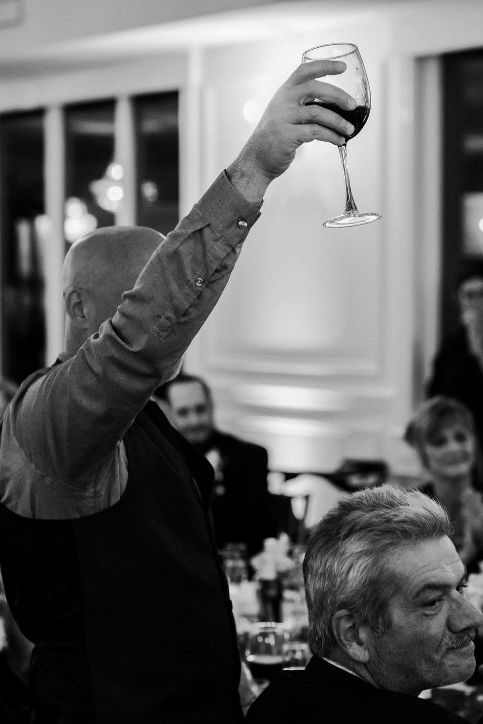 Man holding up a wine glass in a toast at a formal gathering. People seated in the background. Black and white photo.