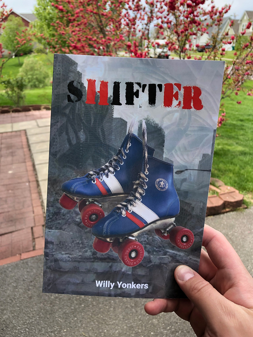 Holding a paperback book with a pair of rollerskates on the cover. Title reads: SHIFTER Author reads: Willy Yonkers.