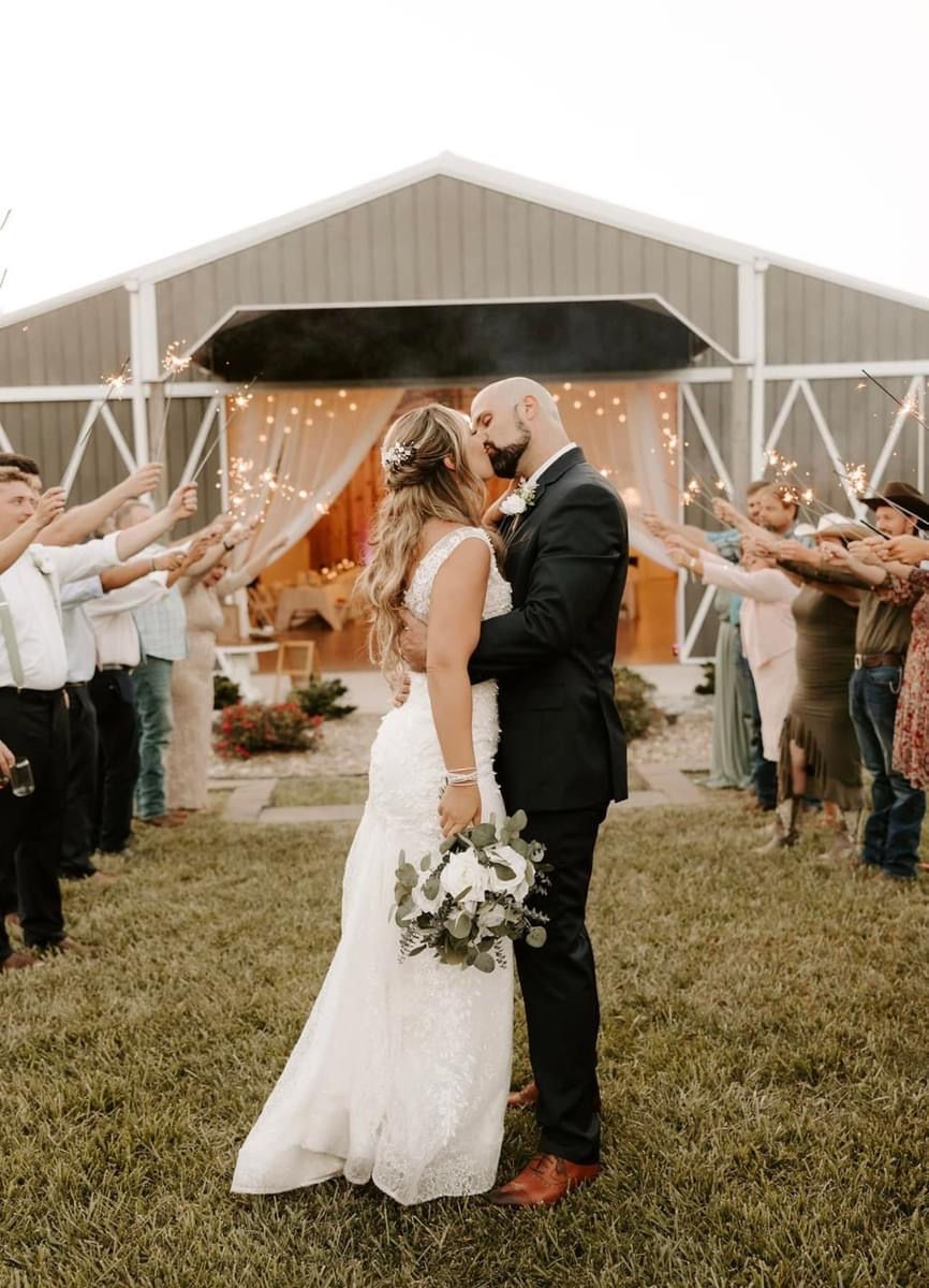 Bride and groom kiss on grass with barn backdrop. Guests hold sparklers, creating a festive and romantic atmosphere.