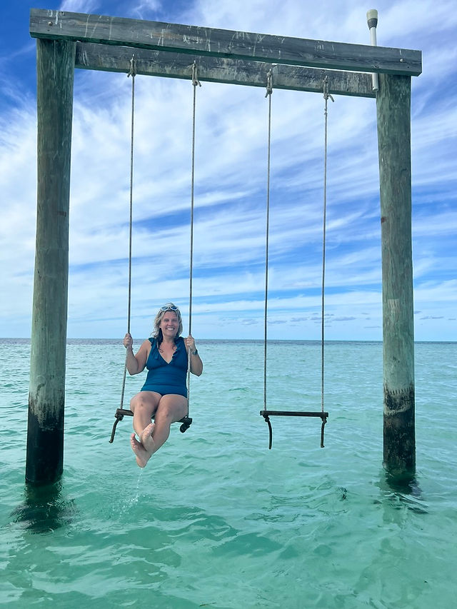 Beautiful woman enjoying the ocean swing, surrounded by crystal-clear blue water, creating a fun and picturesque tropical experience at a sandbar in the Bahamas.