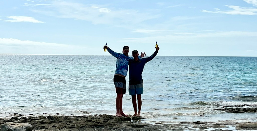 Two good looking brothers standing on a beautiful beach in the Bahamas, holding Bahamian beers with arms outstretched, with the stunning ocean and clear blue sky in the background, enjoying a relaxing tropical vacation.