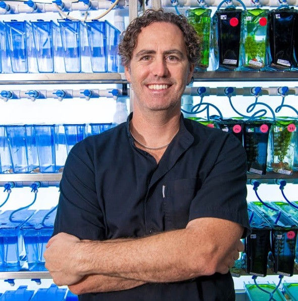 Smiling man with crossed arms stands before rows of blue and green aquarium bags and fish tanks in a bright laboratory