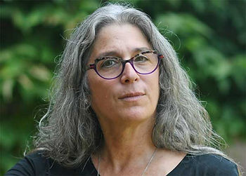 Woman with long gray hair and purple glasses gazes forward. She's outdoors with green foliage in the background, wearing a black top.
