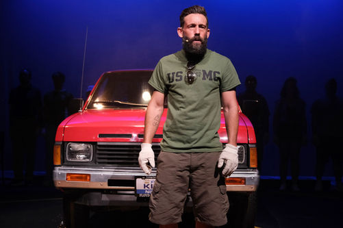 a man with a beard wearing a us marines shirt stands in front of a red truck
