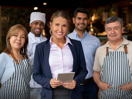 A group of five smiling restaurant team members, including a chef and two in striped aprons, stand confidently in a warmly lit setting. A woman holds a tablet.