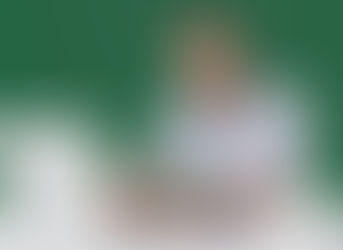 Man with glasses and arm tattoo types on a laptop, smiling. He's in a striped shirt against a green background. A phone and cup nearby.