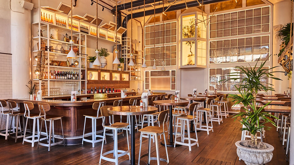 Stylish cafe with wooden tables and bar stools, decorated with green plants and warm lighting. Bottles line the shelves, creating a cozy ambiance.