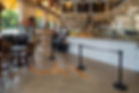 Bright bakery interior with wooden chairs and tables, marble counter displaying pastries. Yellow accents, bread shelf, and cake in background.