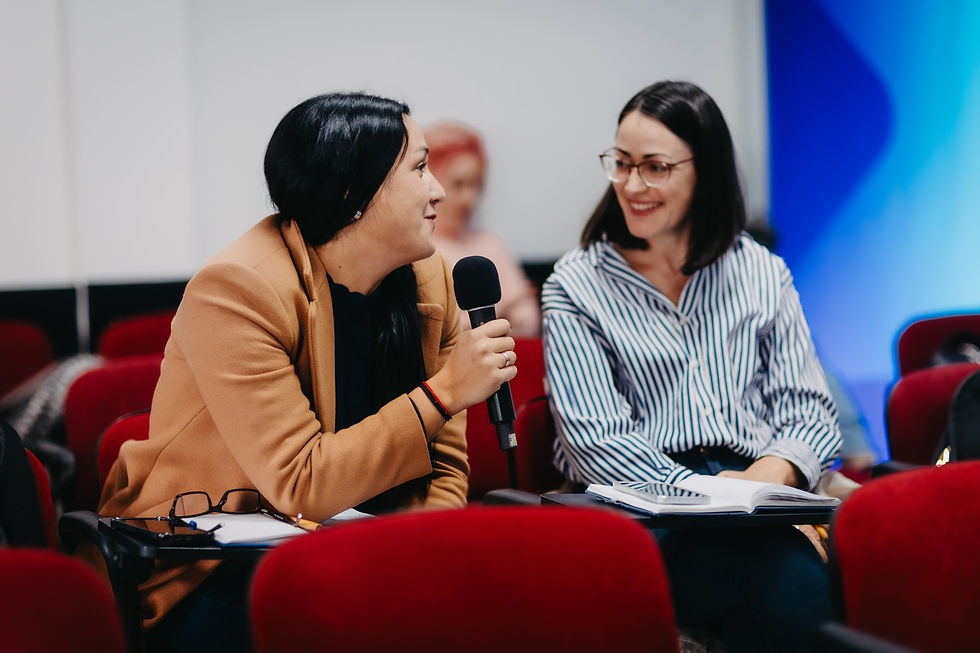Two women sit in red chairs, one holding a microphone, smiling in conversation. Background is blurred, creating a friendly, engaging mood.
