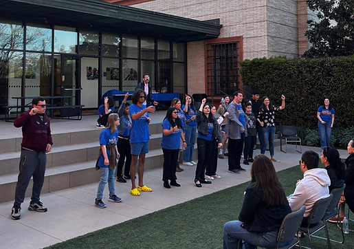 A group of participants in blue Miracle Project shirts standing outdoors performing while in front of a modern building while others are seated in front watching.