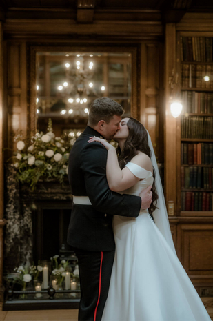 bride and groom kissing, in front of ornate fireplace in old library with lots of wood. Fireplace is dressed with white flowers