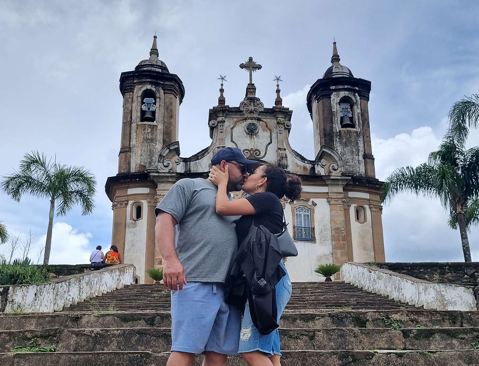 Casal se beija nos degraus de igreja histórica com torres barrocas, cercada por palmeiras sob céu parcialmente nublado.