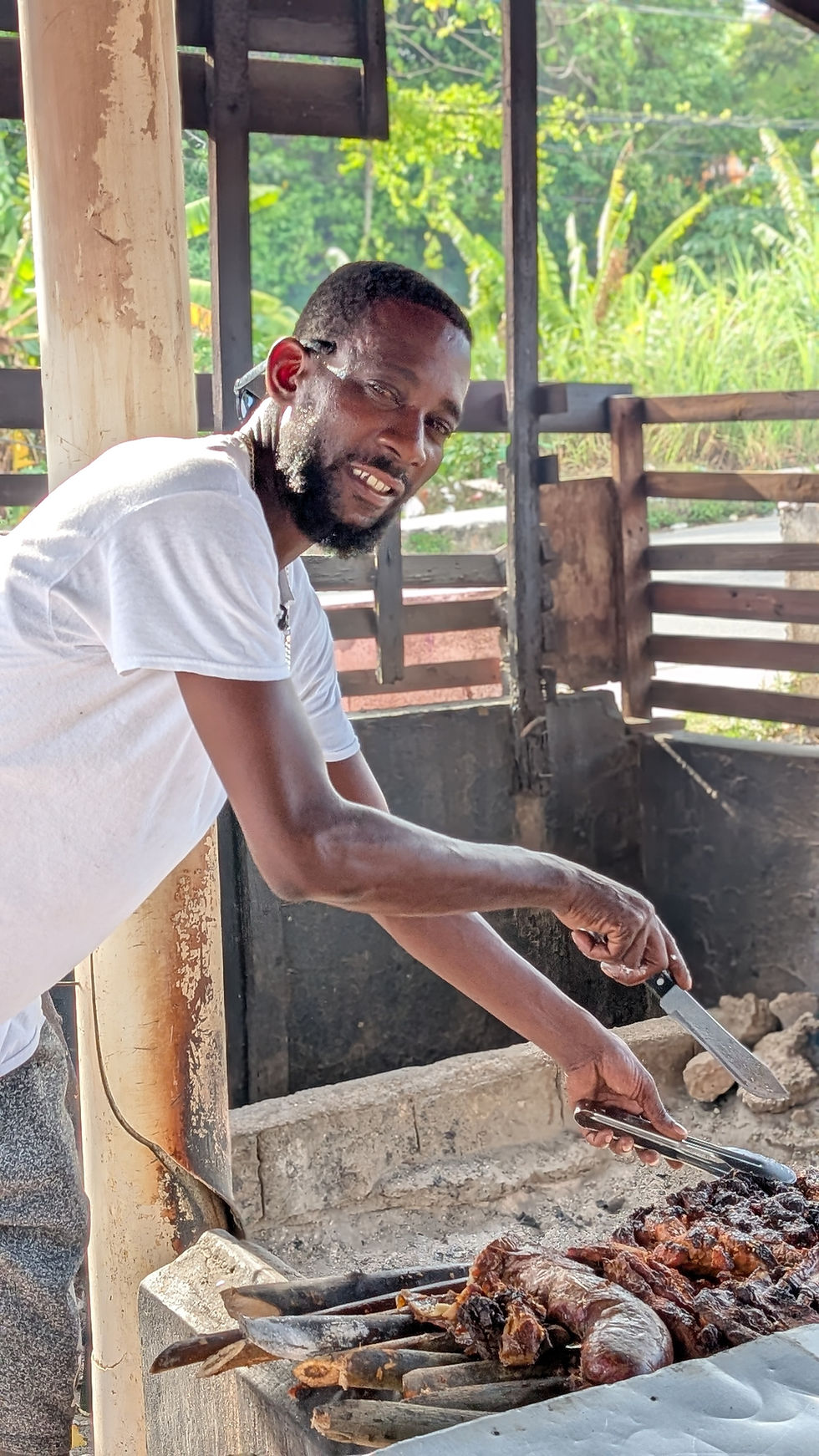 Eye-level view of a rustic outdoor jerk chicken stand with smoke rising and colorful surroundings