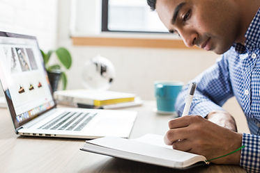 Man with black hair, wearing a blue checkered shirt sits in front of an open laptop. He is using a pen to write in a notebook with his left hand.