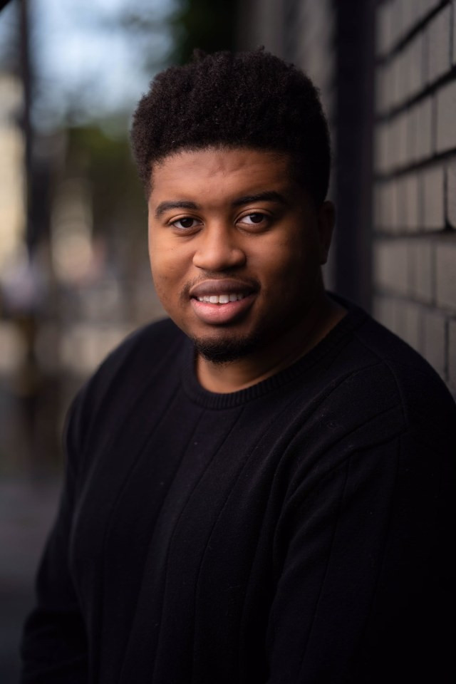 Portrait photo of Dalvin Josias Sejour. He is wearing a black sweater and the photo was taken outside. There is a brick wall behind his left shoulder.
