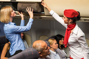 cabin attendant helping passenger stow carry-on bag in overhead bin