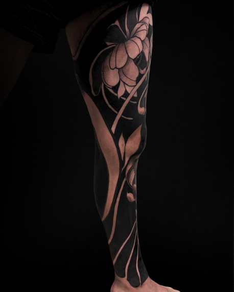 Side angle of a blackout floral leg sleeve tattoo with heavy black saturation, floral elements, and ornamental negative space wrapping around the leg.