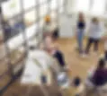 Teammeeting in einem offenen Büro mit Holzboden und großen Fenstern. Mehrere junge Menschen stehen oder sitzen, diskutieren, schreiben auf ein Whiteboard oder nutzen Laptops und Smartphones.
