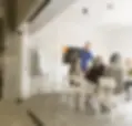 Das Bild zeigt ein Team, das in einem modernen Konferenzraum mit Glaswänden zusammensitzt. Ein Mann steht am Tisch und spricht zu den anderen Personen. Im Hintergrund sind ein Flipchart und ein Bildschirm an der Wand zu sehen.