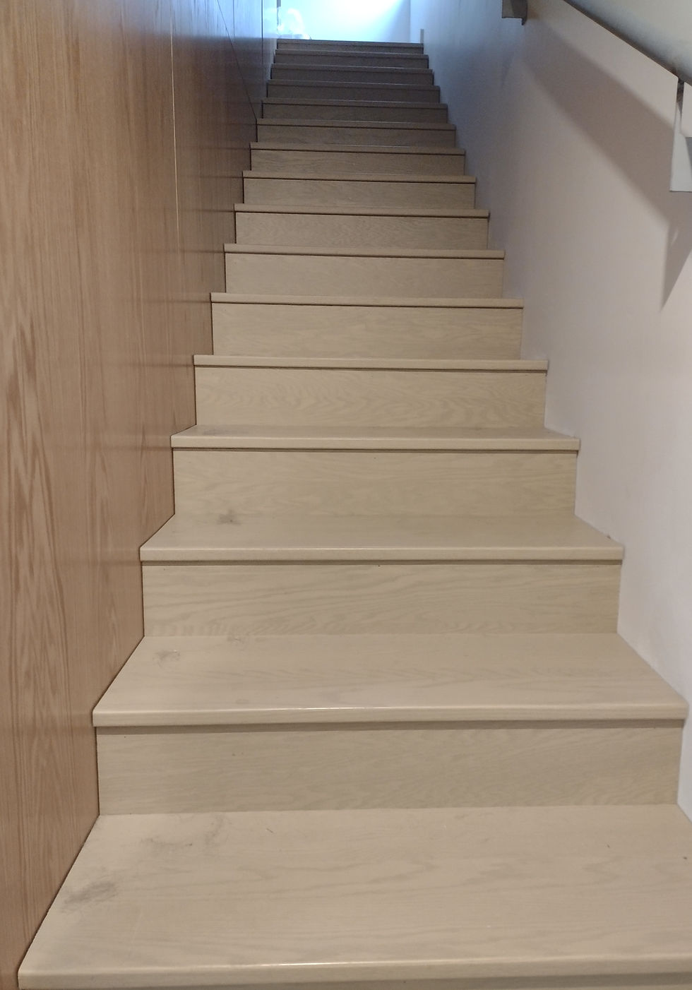 Portrait photo of light wood stairs shot from the bottom looking up, walls on both sides, bright opening at the top, neutral tones, strong vertical lines, center stairs, portrait aspect ratio.