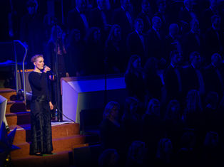 A photograph of Rachael Heater, soprano, singing from the choir stalls in the Waterfront Hall, lit by a single spotlight.