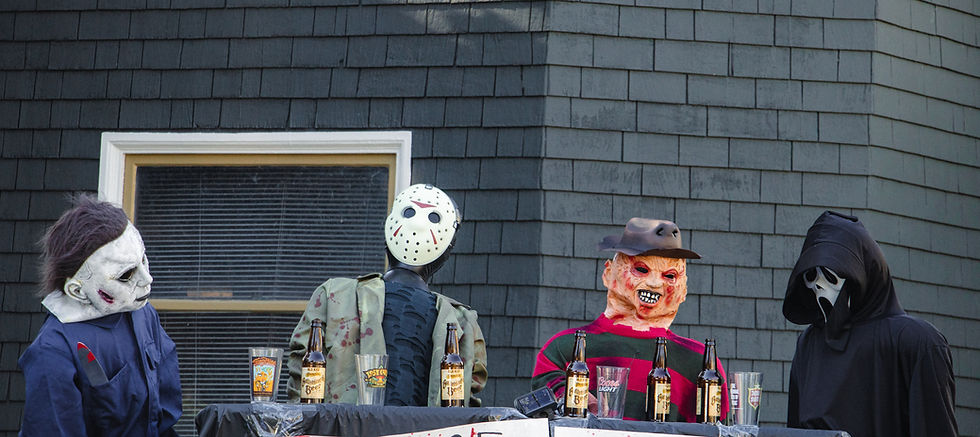 Four horror character mannequins sitting at a table with beer bottles against a gray shingle wall. Creepy, casual atmosphere.