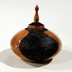 A stout, rounded wooden container with a delicately carved pointed top and inky black decorative texture.