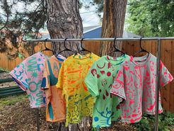 Five brightly colored, patterned screen-printed shirts hanging on a bar, with a tree in the background.