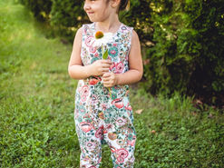 A smiling child in nature, dressed in a florally patterned romper and holding a flower.