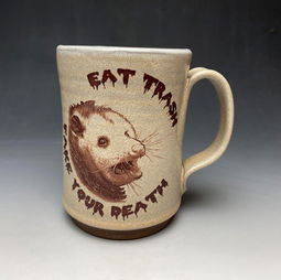 A tan ceramic mug depicting the face of a possum with the caption, "EAT TRASH, FAKE YOUR DEATH."
