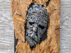 A molten metal mask encircled by wood and nails on a black block base.