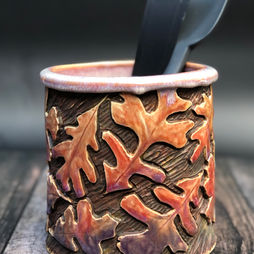 A cylindrical clay vessel, carved and colored into a pattern of orange and purple leaves against a textured brown base.