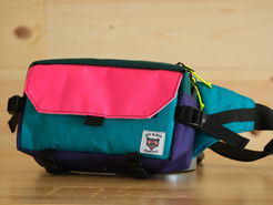 A bright pink, blue and purple fanny pack with black trim and a small tag reading, "Sew Bueno Company."